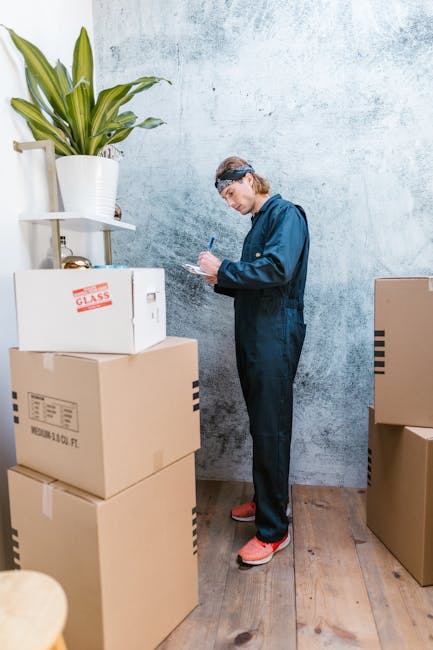 A person dressed in a blue work jumpsuit and red footwear standing inside an indoor space with a textured, light grey wall. They are holding a pen and a clipboard, appearing to record details during a